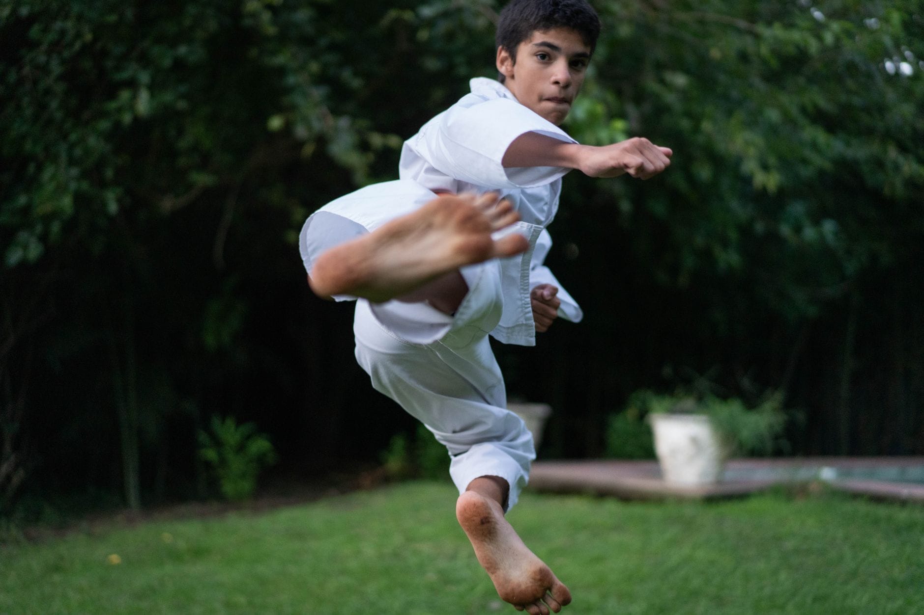 Nastolatek w białym gi ubrany do jujutsu wykonuje wysoki kopniak w ogrodzie
